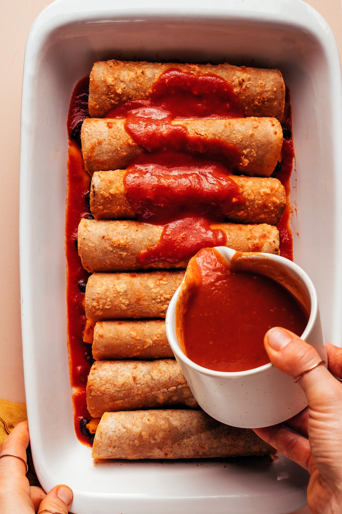 Pouring enchilada sauce over rolled up tortillas stuffed with roasted butternut squash and black beans
